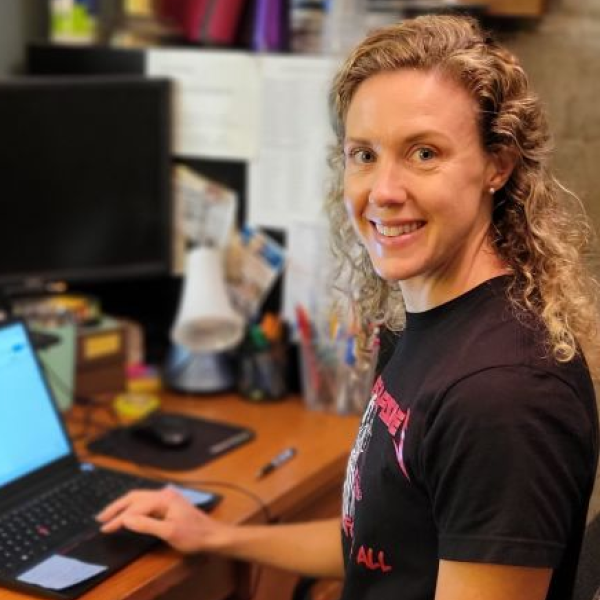 Kate Fisher, woman with medium to long blond curly hair is sitting with a laptop on a table.