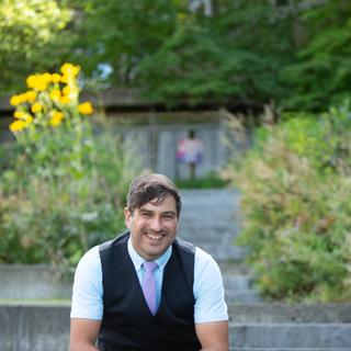 Michael MacKenzie, man with short brown hair, is sitting outside in front of garden and treed area